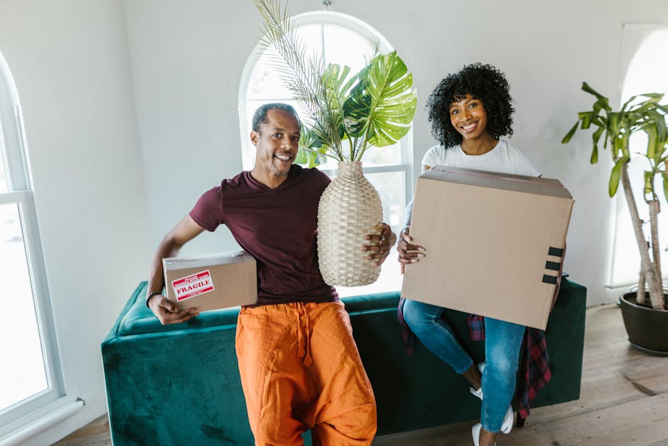 A man and a woman are inside a bright, modern living room with white walls and large arched windows, preparing for a home relocation. The man, dressed in a maroon t-shirt and orange trousers, is smili